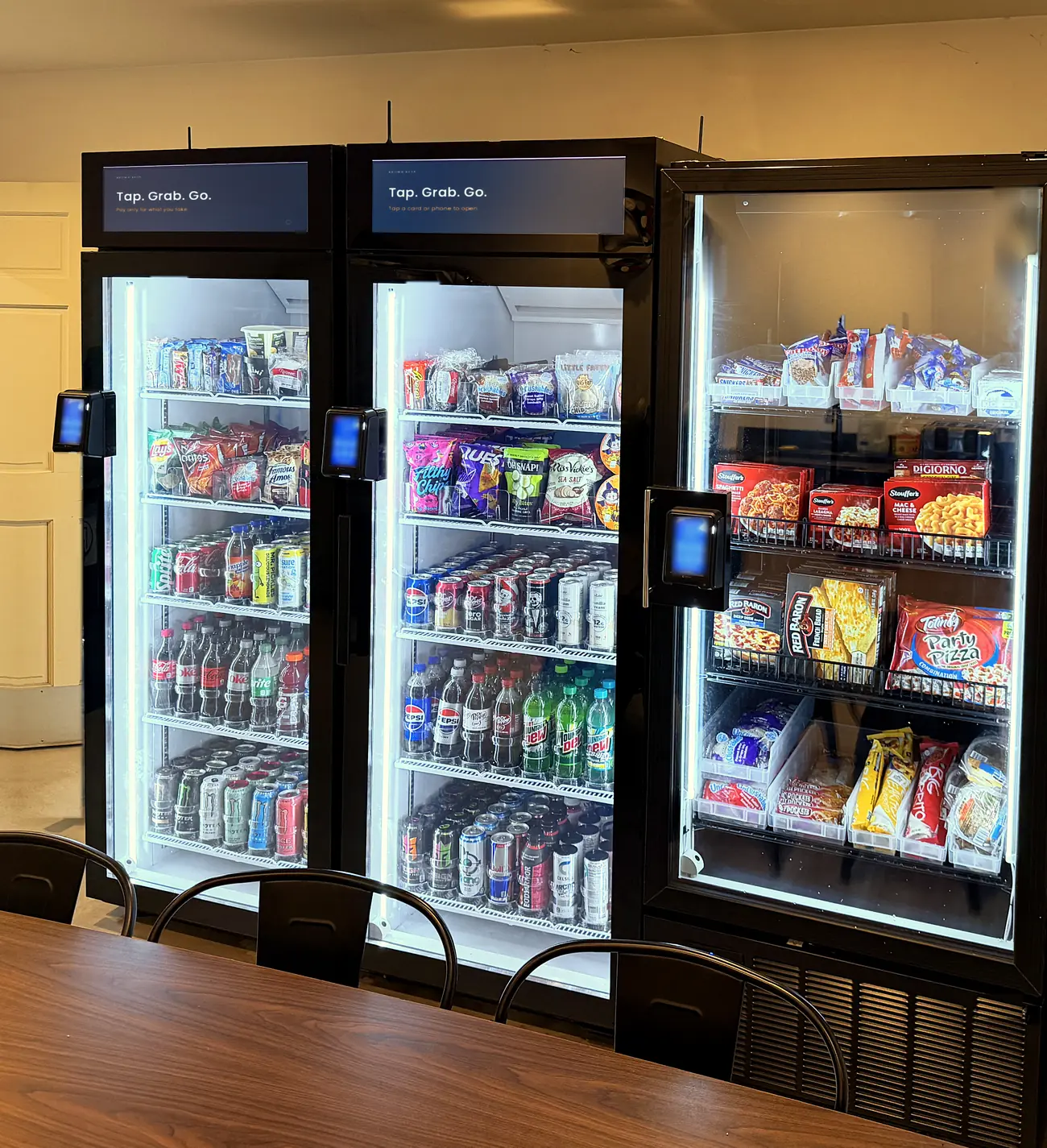 A bright break room with three Brown Bear smart coolers, lit and fully stocked, behind a wooden table and chairs.
