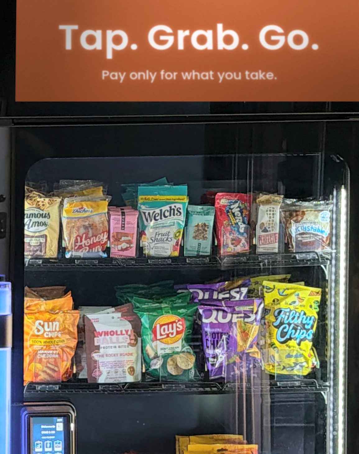 A Brown Bear smart cooler with a Tap. Grab. Go. screen on top, snacks on the shelves and a tap-to-pay reader on the door.