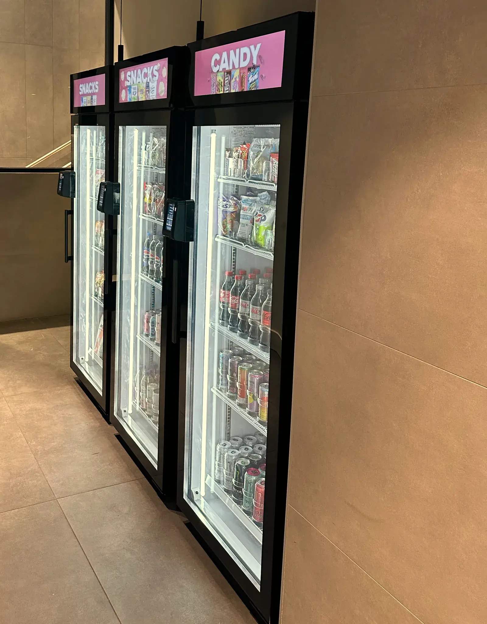 Three Brown Bear smart coolers in a quiet lobby alcove, lit and stocked, with Snacks and Candy screens on top and a tap-to-pay reader on each door.