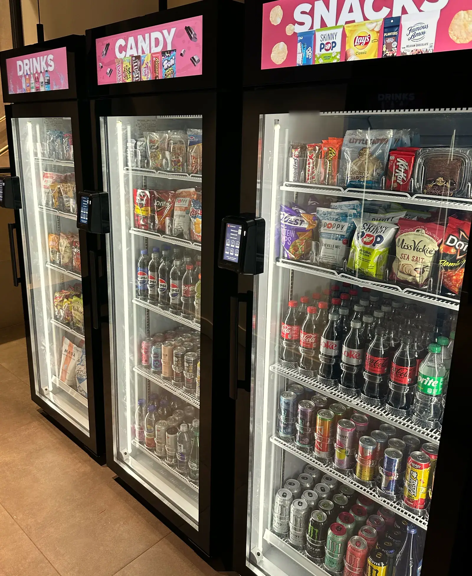 Three Brown Bear smart coolers side by side, lit and fully stocked, with Drinks, Candy and Snacks screens on top and a tap-to-pay reader on each door.
