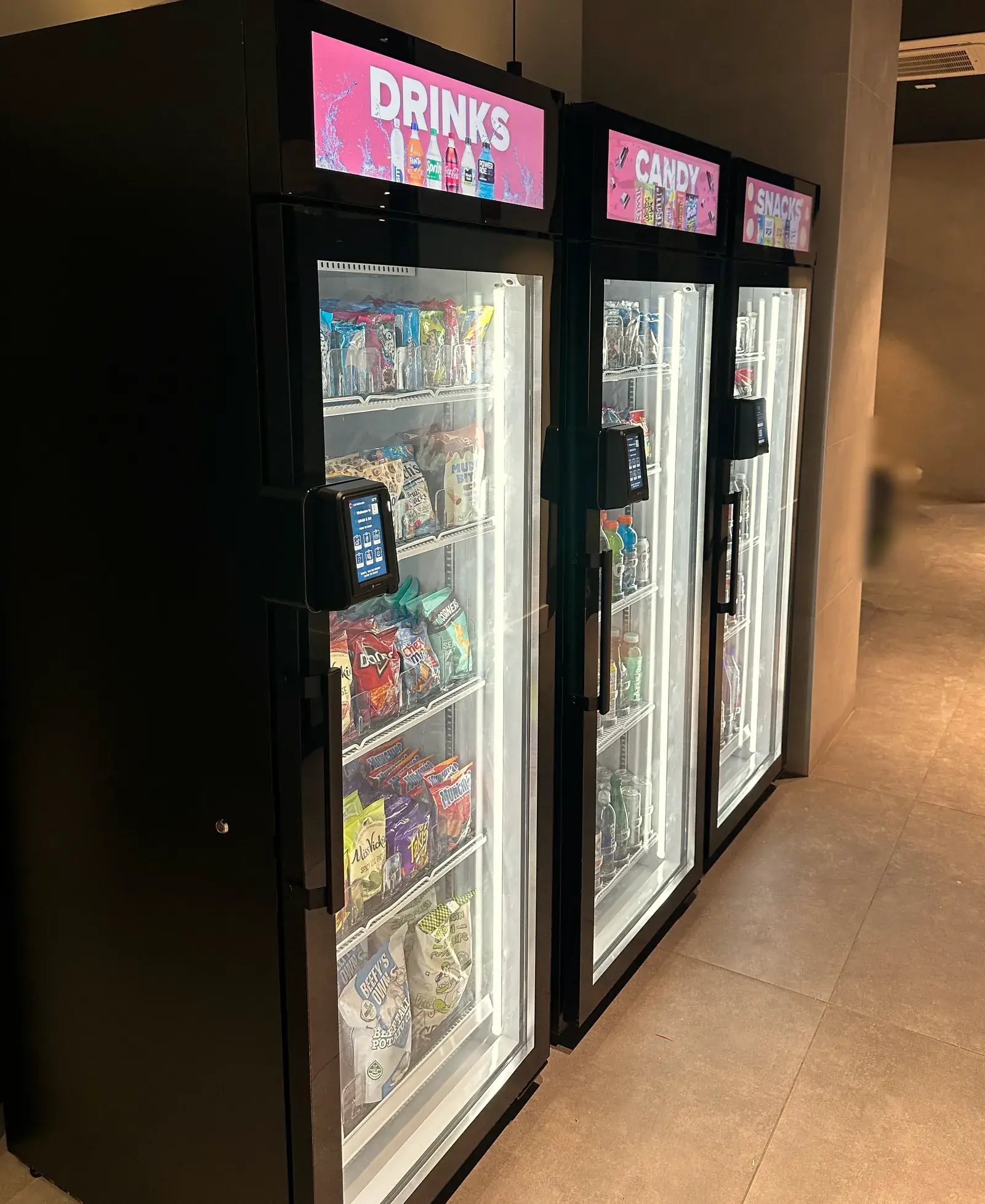 A row of three Brown Bear smart coolers along a hallway, lit and stocked, with Drinks, Candy and Snacks screens on top.
