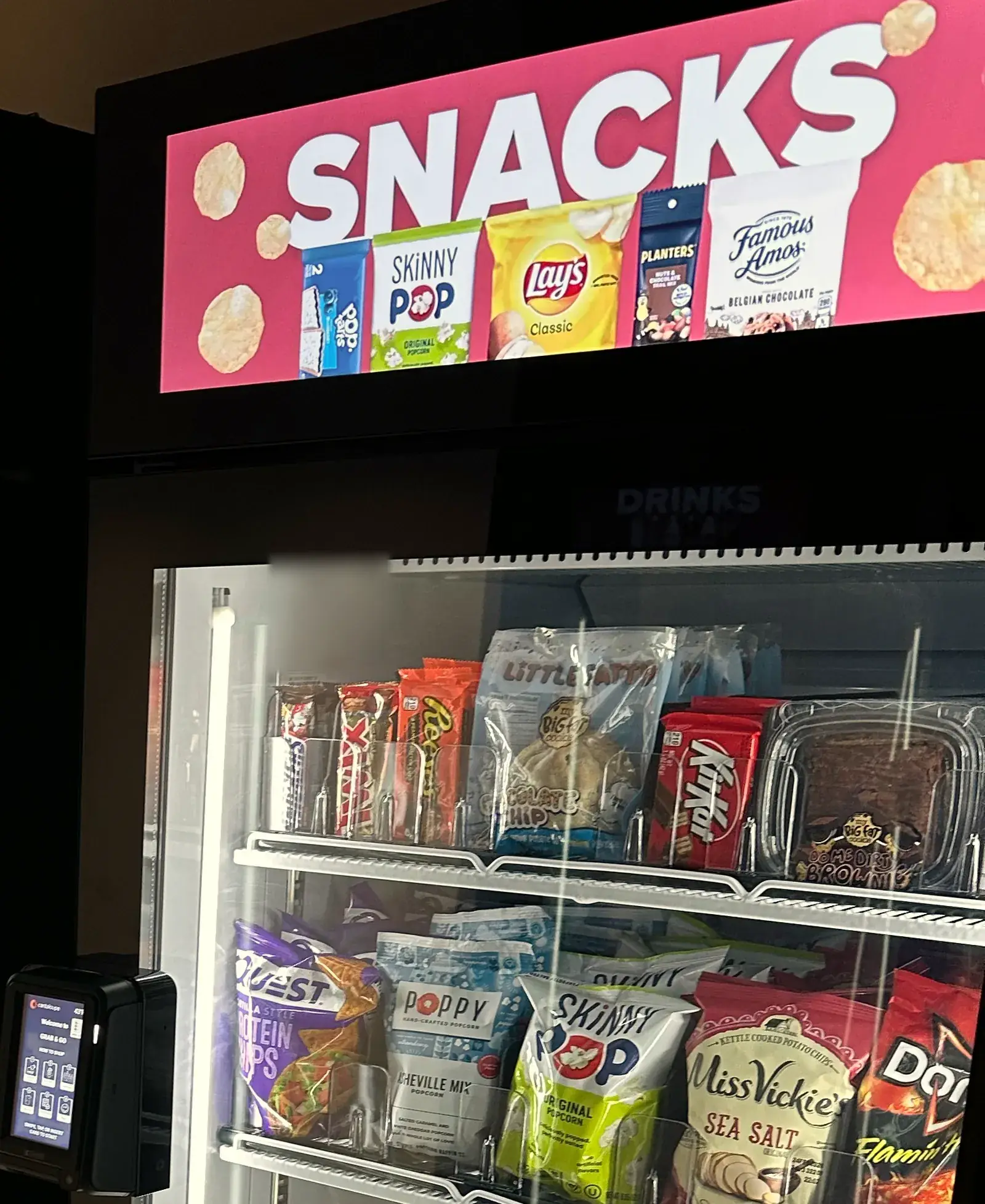 A Brown Bear smart cooler with a bright Snacks screen on top, stocked with chips, candy and snack packs behind the glass door.