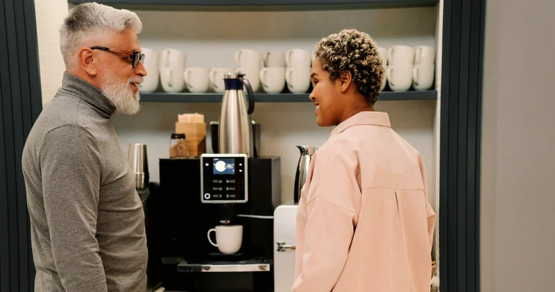 Two colleagues chatting by a bean-to-cup coffee machine in an office break room.