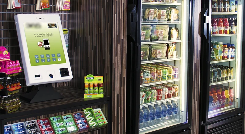 A micro-market with a pay kiosk, snack shelves and glass-door coolers of fresh food and drinks.