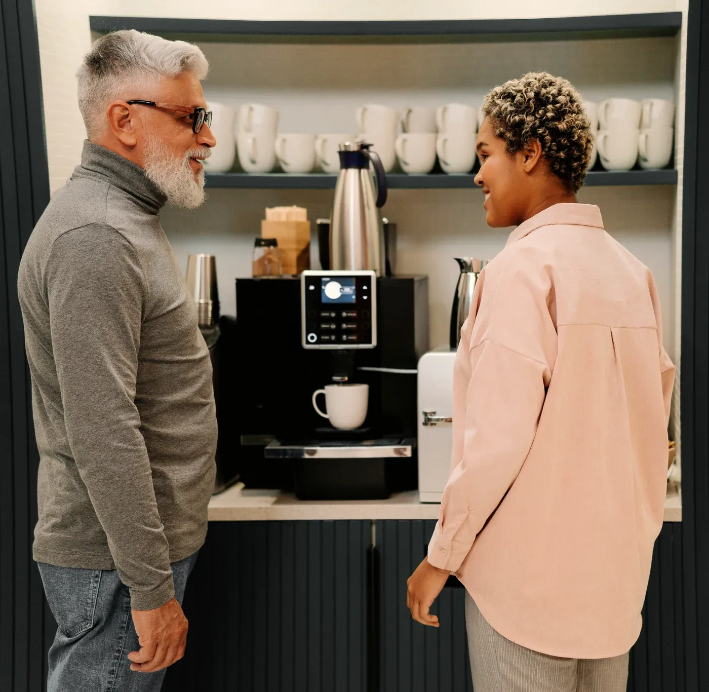 Two colleagues chatting by a bean-to-cup coffee machine in an office break room.