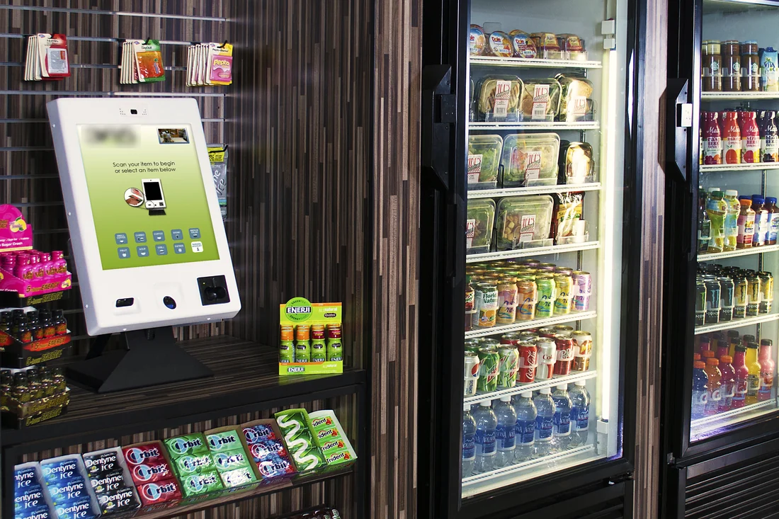 A micro-market: a pay kiosk on wood shelving stocked with snacks, beside glass-door coolers of fresh salads, sandwiches and cold drinks.