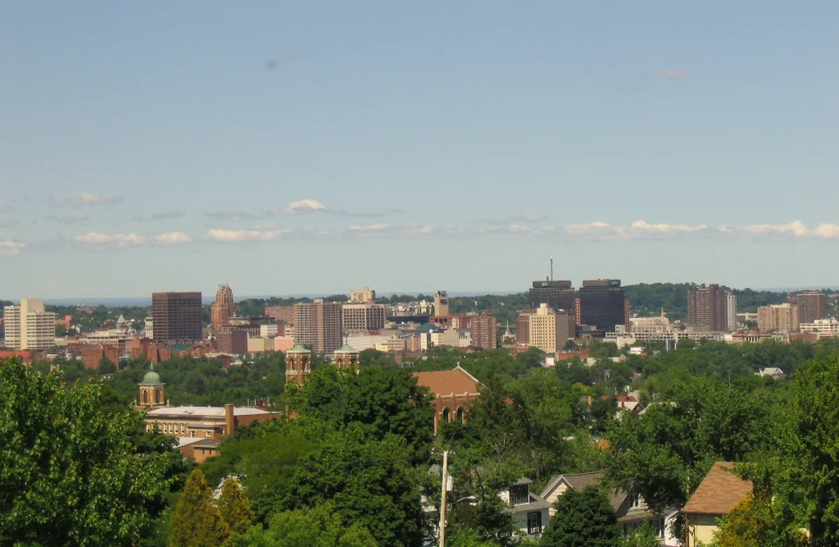 The downtown Syracuse skyline above green treetops on a clear day.